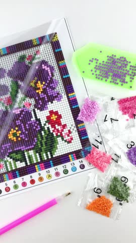 Assembled purple flower diamond mosaic fragment, tray with rhinestones, pen tool, rhinestones in transparent packaging and folded protective film, white background, close-up, top view, vertical, DIY