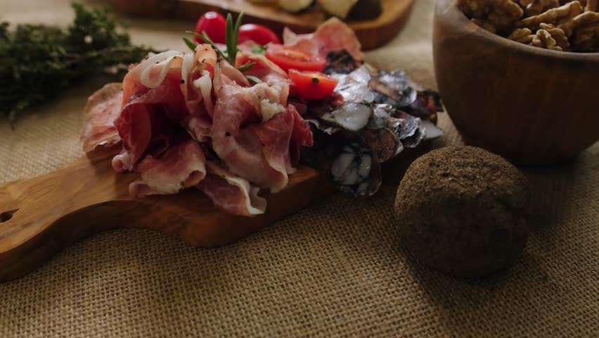 Close-up still life of cheese and meat delicacies. Craft cheeses and cold meat are on the table among aromatic rosemary, cherry tomatoes and other snacks. Gourmet plate antipasto setting. Cheese board