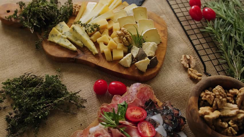 Close-up still life of cheese and meat delicacies. Craft cheeses and cold meat are on the table among aromatic rosemary, cherry tomatoes and other snacks. Gourmet plate antipasto setting. Cheese board