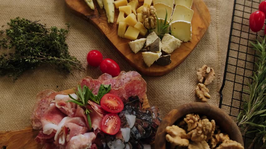 Close-up still life of cheese and meat delicacies. Craft cheeses and cold meat are on the table among aromatic rosemary, cherry tomatoes and other snacks. Gourmet plate antipasto setting. Cheese board