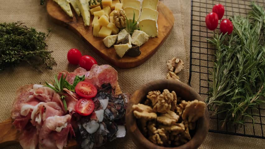 Close-up still life of cheese and meat delicacies. Craft cheeses and cold meat are on the table among aromatic rosemary, cherry tomatoes and other snacks. Gourmet plate antipasto setting. Cheese board