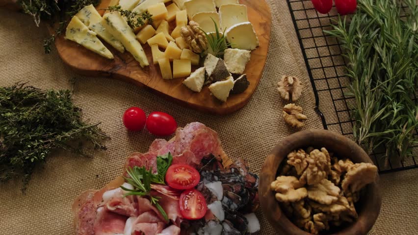 Close-up still life of cheese and meat delicacies. Craft cheeses and cold meat are on the table among aromatic rosemary, cherry tomatoes and other snacks. Gourmet plate antipasto setting. Cheese board