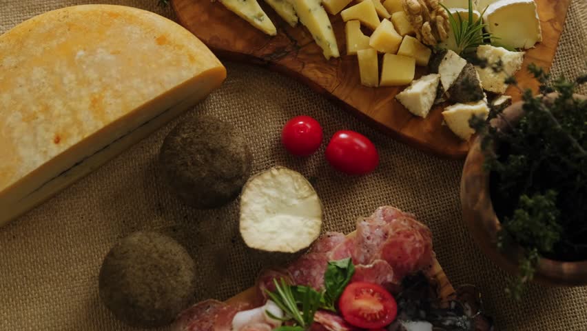 Close-up still life of cheese and meat delicacies. Craft cheeses and cold meat are on the table among aromatic rosemary, cherry tomatoes and other snacks. Gourmet plate antipasto setting. Cheese board