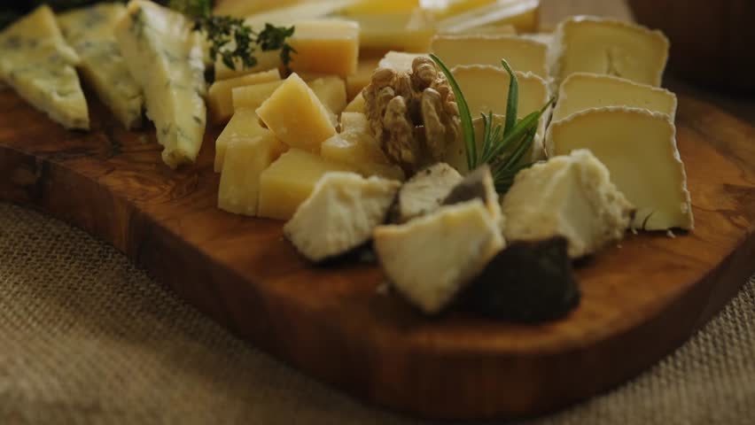 Close-up still life of cheese and meat delicacies. Craft cheeses and cold meat are on the table among aromatic rosemary, cherry tomatoes and other snacks. Gourmet plate antipasto setting. Cheese board