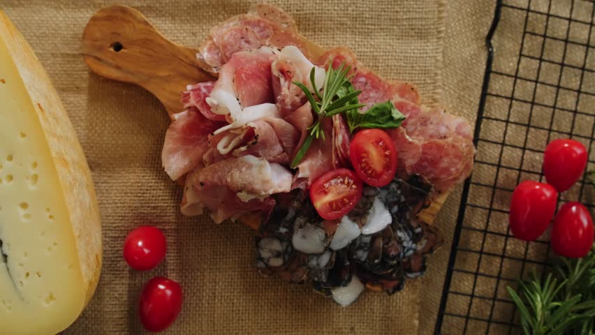 Close-up still life of cheese and meat delicacies. Craft cheeses and cold meat are on the table among aromatic rosemary, cherry tomatoes and other snacks. Gourmet plate antipasto setting. Cheese board