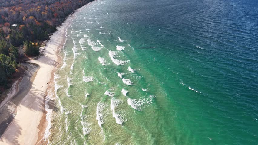 Drone shot of waves rolling onto a sandy beach backed by autumn-colored forest in Michigan’s Upper Peninsula