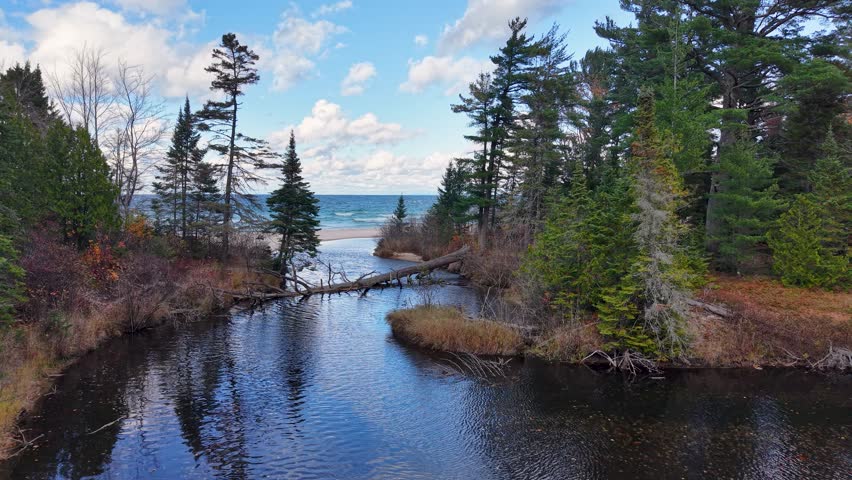 Aerial drone view of a narrow river opening into Lake Superior, framed by evergreens and calm reflections in Michigan’s Upper Peninsula