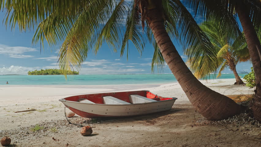Small red and white boat resting on a pristine white sand beach under the shade of palm trees in Aitutaki, Cook Islands, with turquoise waters and a small island in the background