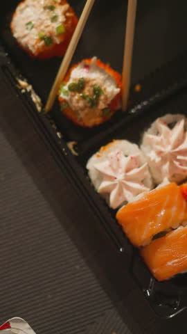 Taking sushi roll with cream cheese cap by wooden chopsticks from plastic tray closeup. Traditional Asian food for dinner at home. Healthy oriental seafood