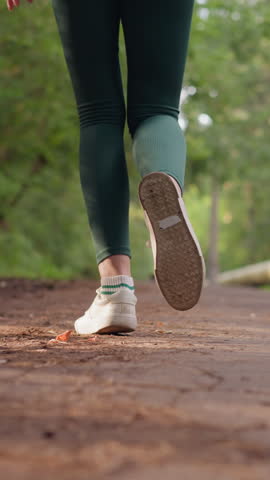 Female jogger running along path in park. Woman legs in sports shoes running on pathway surrounded by trees. Lady training in nature on summer day