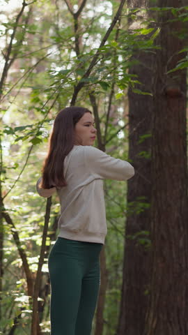 Young woman athlete stretches arms in forest. Playing sports surrounded by different types of trees in wild in sunny weather. Sun rays fall on lady