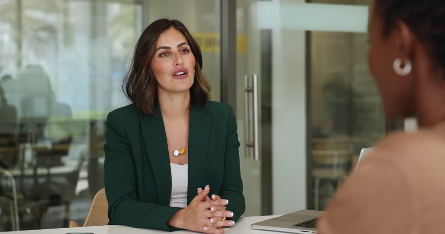 Confident Hispanic young professional woman talking to female African colleague, discussing partnership, teamwork, hiring, employment terms, giving handshake over meeting table