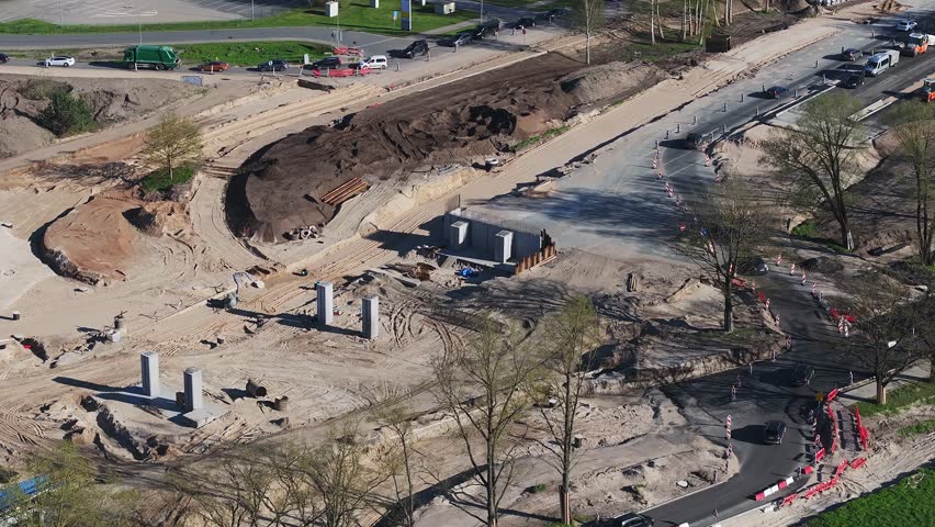 Aerial view of an active construction site in Riga, Latvia, showing workers, heavy machinery, concrete pillars, and nearby roads with light traffic.