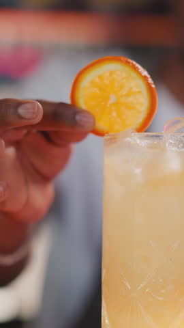 Bartender puts orange slice and straw into cocktail closeup. African American man decorates citrus beverage in nightclub. Refreshing party drink