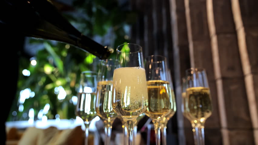 Pouring champagne into multiple glasses at a festive celebration, close-up view of sparkling wine being served at a luxury event with green plants and blurred background
