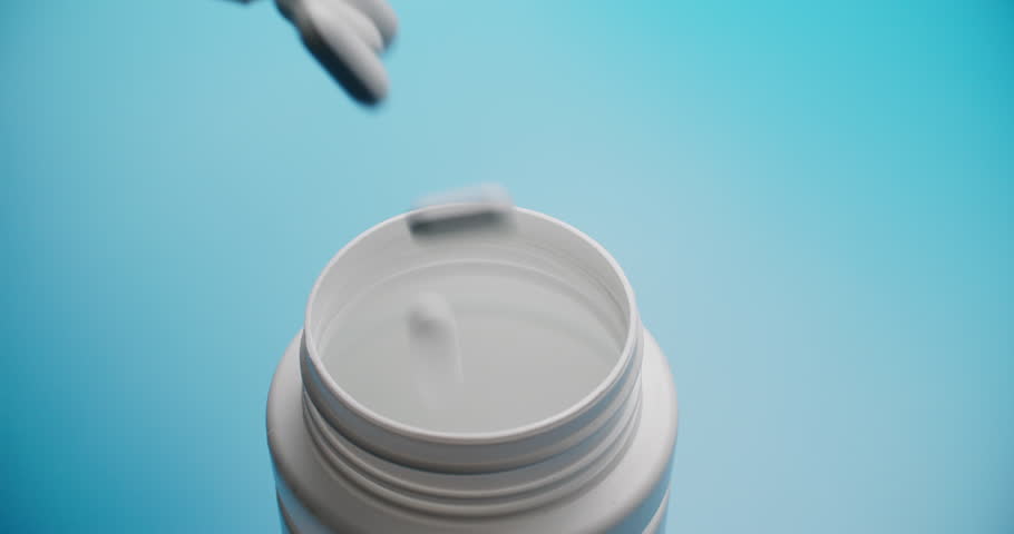 Slow motion close-up of a white pill bottle against blue background with grey tablets falling in foreground. Soft focus creates dreamy medical aesthetic with strong color contrast.