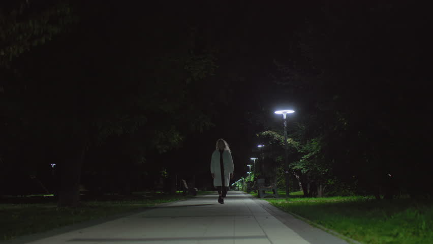 Young lady wearing white coat strolls alone in quiet park at night, subtle presence trailing behind her under soft lamp glow, capturing calm mood yet hint of tension, trees shadowy