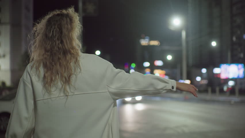 Young woman extends arm to stop taxi under streetlights while walking toward parked white car on roadside in nighttime urban setting with blurred city lights and passing vehicle in background