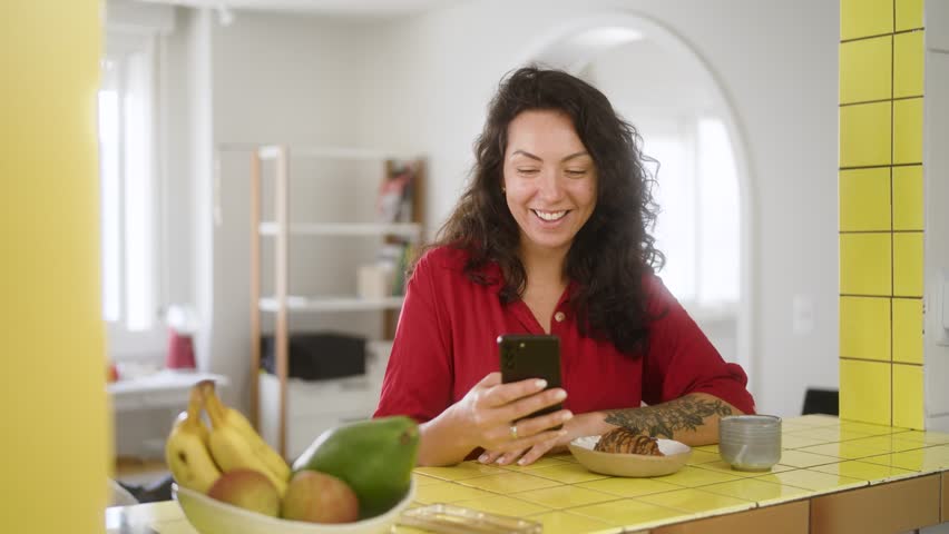 Smiling Latin woman enjoying a chocolate croissant while sipping coffee in a vibrant kitchen. Cheerful moment using a mobile phone during a relaxed morning.