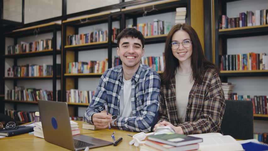 Two students smiling while studying together in a library filled with books during daytime