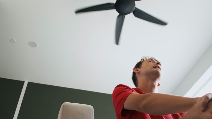 Man sitting under ceiling fan at home in living room, cooling off during hot summer weather, suffering from heat, high temperature. Sweaty exhausted male lifting his red shirt to get cool
