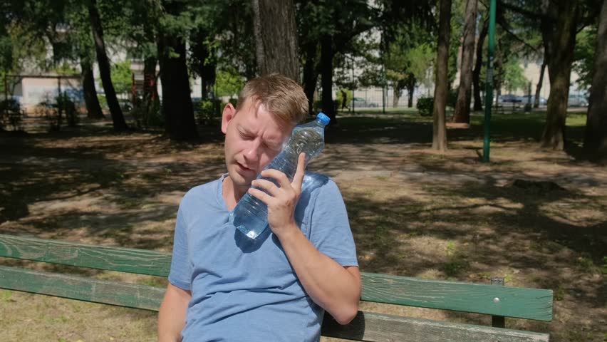 Man suffering from heat wave, guy with heatstroke. Caucasian male presses bottle of water to face to cool off. Exhausted tired overheated man. Sunstroke at summer hot weather