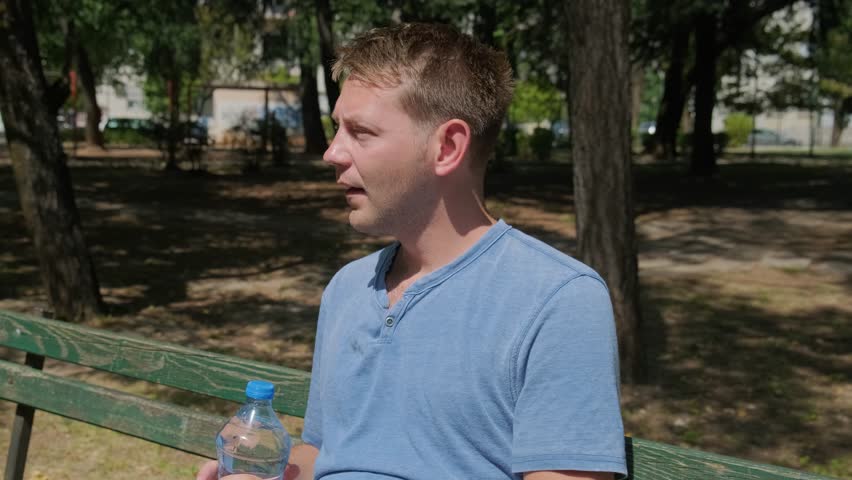 Man suffering from heat wave. Caucasian male drinking water to cool off and get rid of thirst while sitting outdoors. Exhausted tired overheated man sitting on the bench outdoor in the city