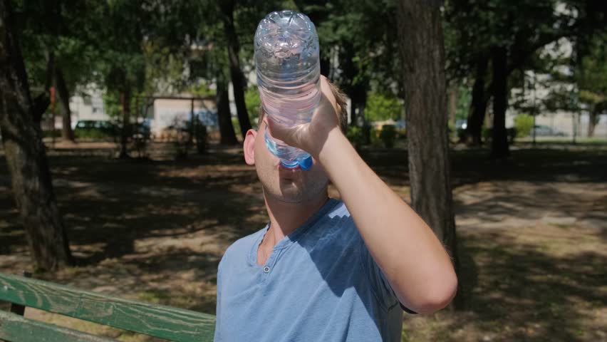 Man suffering from heat wave. Caucasian male drinking water to cool off and get rid of thirst while sitting outdoors. Exhausted tired overheated man sitting on the bench outdoor in the city