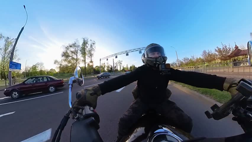 Handlebar cam captures V-twin cruiser leaning through a city intersection. Traffic lights, road markings, greenery visible. Motion blur highlights dynamic turn.