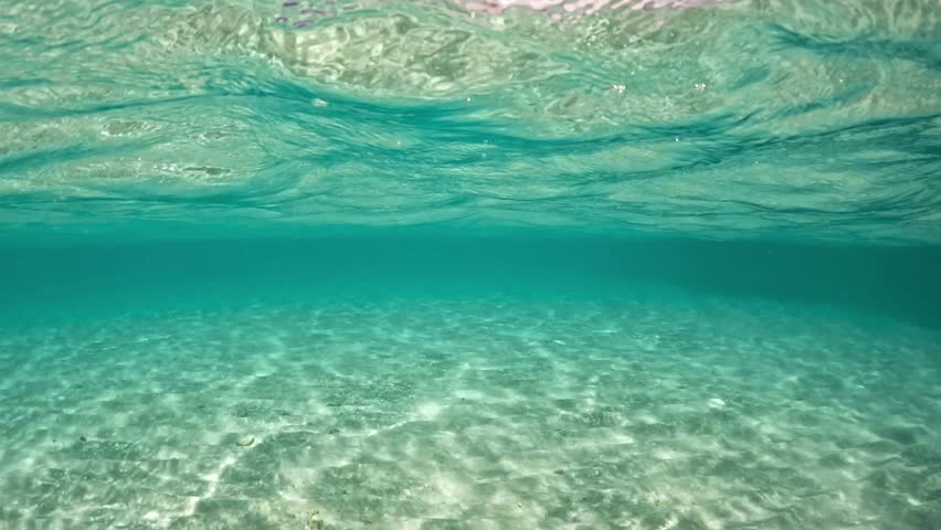 Clear turquoise water above the underwater scenery of Bora Bora, with bright sunlight illuminating the surface and clouds in the sky above.