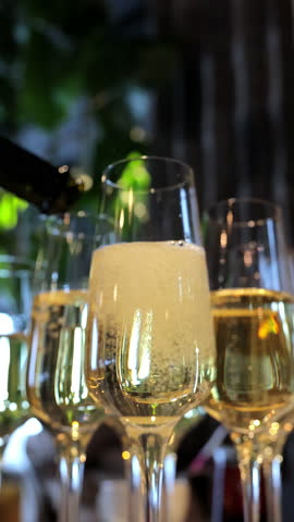 Elegant champagne being poured into flutes at a sophisticated event, detailed close-up of sparkling wine filling glasses with a lush green background and festive atmosphere. Vertical video