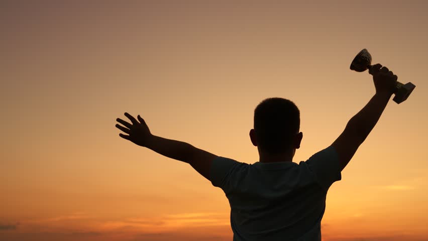 Childhood dream victory. Child with winner cup raises hand rejoices in sport victory outdoor. Best of best child, victory, achievement of success. Winner child, silhouette. Child athlete at sunset sky
