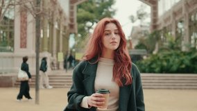 A young woman with long, red hair smiles at the camera while holding a coffee cup in a park. People are walking in the background. - Powered by Shutterstock - Get 15% off with code: PIKWIZARD15