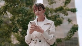 A young businesswoman checks her phone while standing in front of an office building on a sunny day in the city. She smiles at the messages she receives. - Powered by Shutterstock - Get 15% off with code: PIKWIZARD15