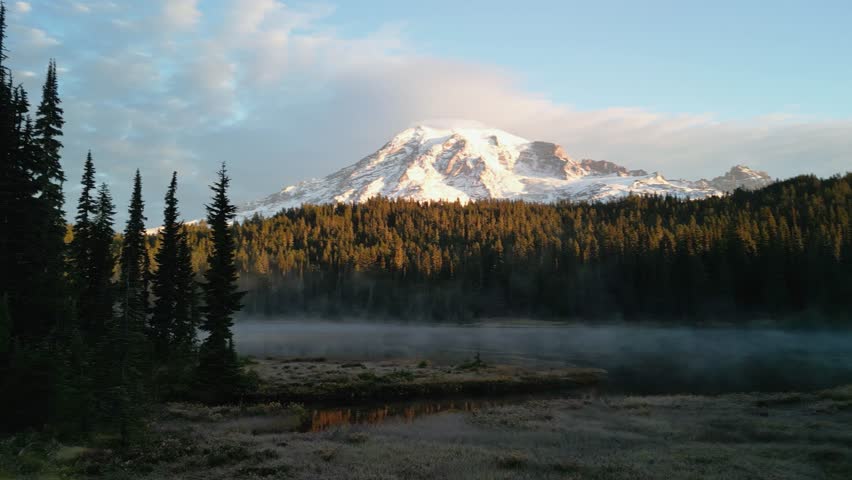 Aerial drone view of Mount Rainier at sunrise, with fog hovering over a calm forest lake in the Pacific Northwest