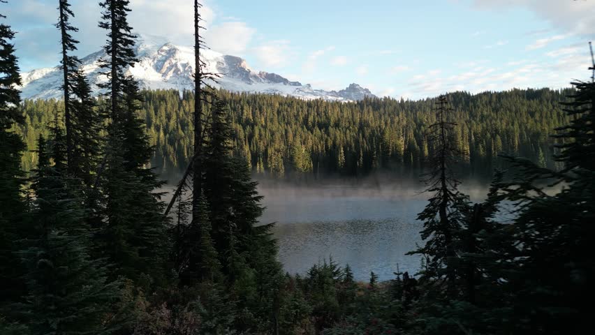 Drone footage of a serene mountain lake surrounded by dense evergreen forest with snowcapped peaks in the distance, Mt. Rainier, Washington