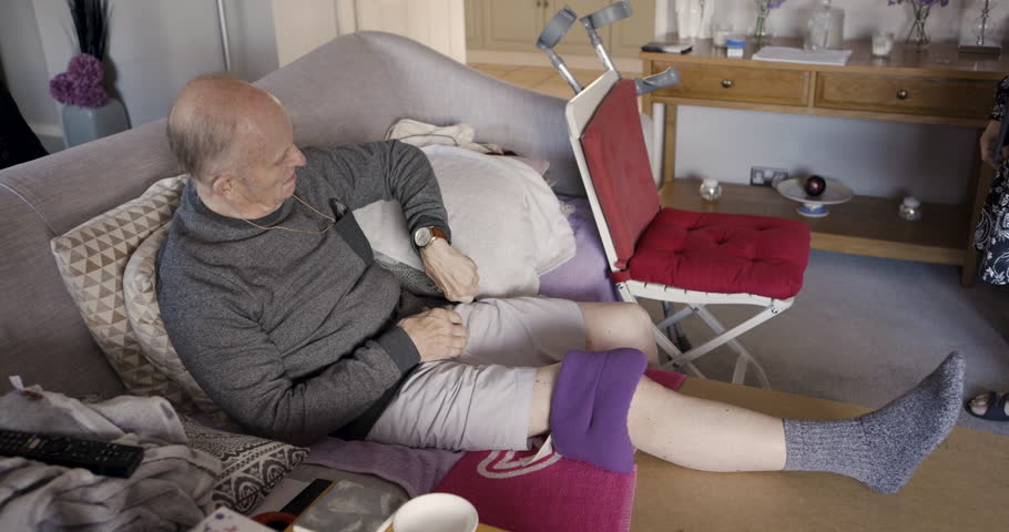 Senior man, talk and resting leg, injury or pain after knee surgery with rehabilitation on sofa in home. Elderly male relaxing, sitting or break in retirement with a bean bag, inflammation or bruise