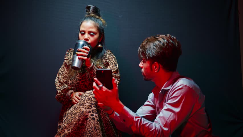 Cameraman leans towards model showing style plan via phone, Coffee in hand lady nods her head at wardrobe idea, Shooter guides with fashion screen shot, Expert holds outfit while girl admires a dress
