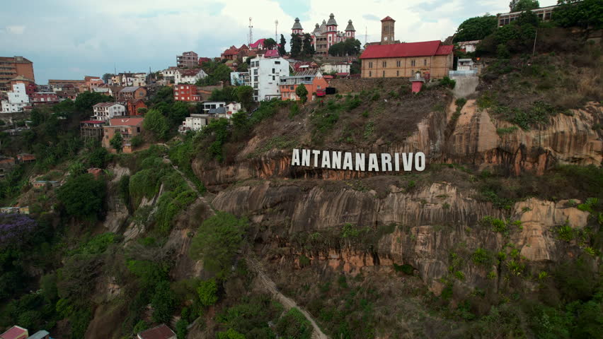 Slow aerial ascent and pullback unveiling the Antananarivo sign, rocky cliffs, and vibrant hillside neighborhoods of Madagascar’s highland capital