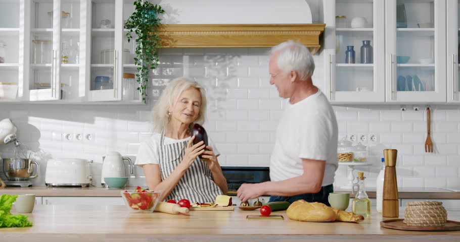 Aged playful married couple dancing in kitchen, portrait of funny grandparents. Elderly spouses listening to favorite song and moving by rhythm, joyful grandma and grandpa cooking dinner at home