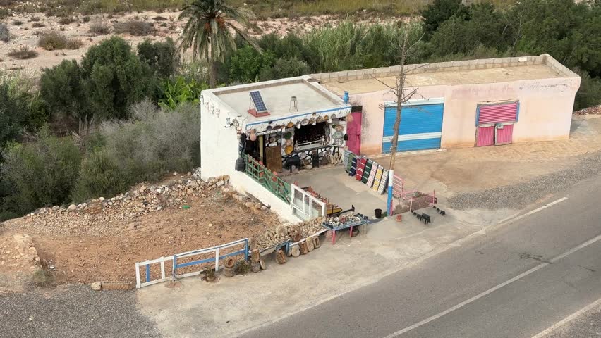 A rustic roadside shop near Agadir, Morocco, with colorful textiles, pottery, and solar panels on display. Surrounded by trees and a dirt lot, it highlights rural charm and sustainability.