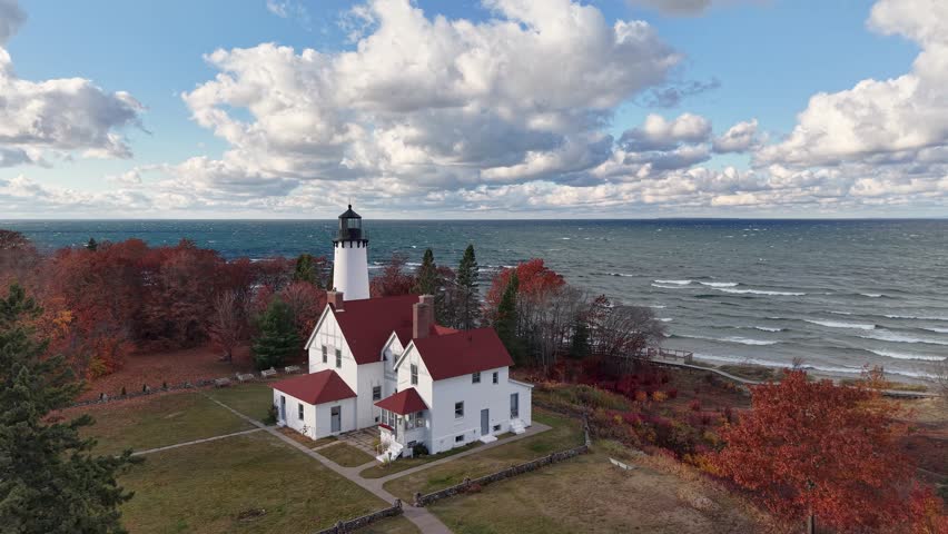 Aerial drone shot of a historic white lighthouse on Lake Michigan’s shoreline, surrounded by autumn trees and crashing waves