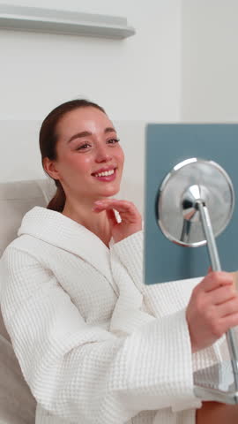 Caucasian girl sitting in beauty clinic and smiling while checking face in mirror after cosmetology procedure. Indian young woman feels satisfied with treatment, enjoying healthy fresh skin result.