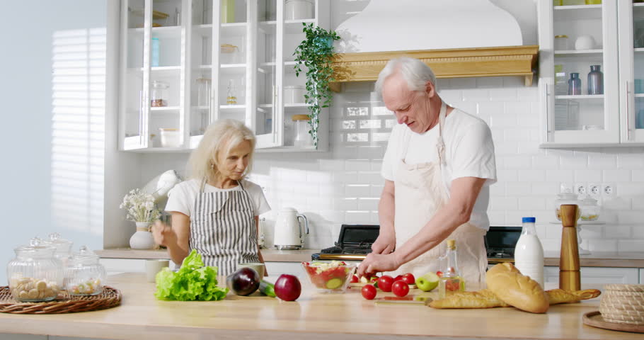 Healthy food and nutrition, aged man cooking salad for pretty elderly wife. Old married couple spending weekend morning in home kitchen, aged lady enjoying coffee when her husband cutting vegetables