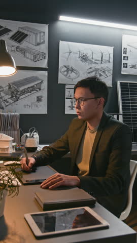 Vertical side shot of male Asian engineer designing sustainable technologies, drawing on art tablet and looking at computer, working in modern renewable energy office