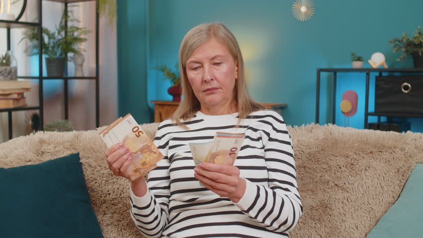 Happy Caucasian mature woman counts money euro cash in hands after winning lottery bonus or receiving a gift. Grandmother smiles broadly sitting on home sofa, celebrating financial victory salary