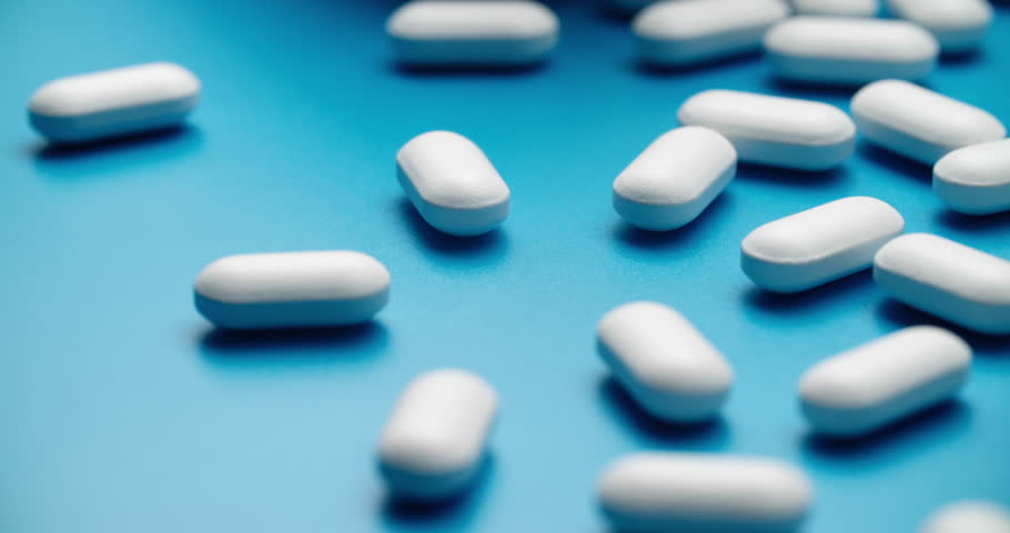 Slide close-up of white pills spilling from a bottle held by hand against solid blue background. Dynamic medical scene with scattered falling tablets.