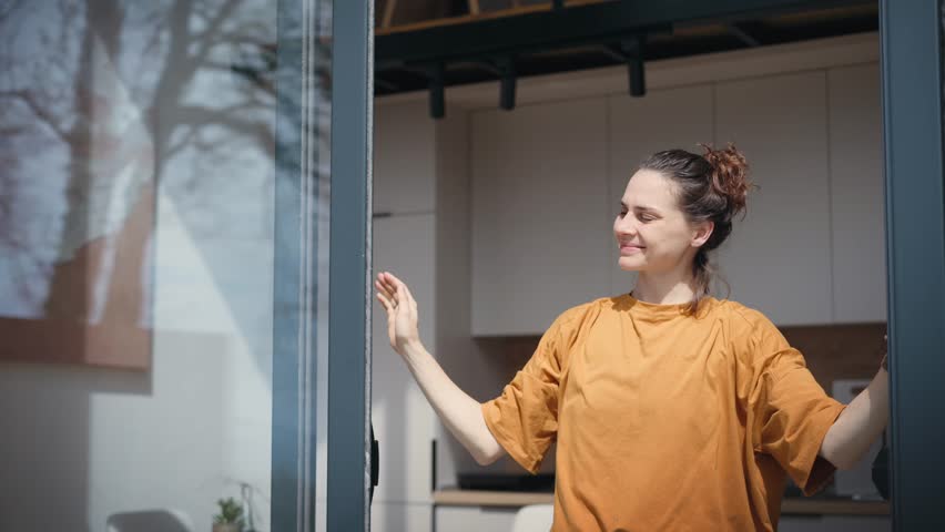 A young Caucasian woman enjoys the view while standing by the panoramic window, against the background of a bright kitchen