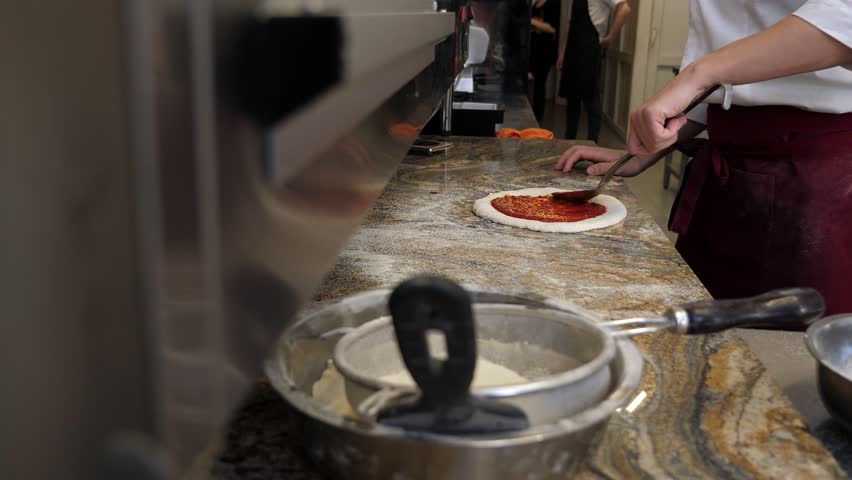 Cook or baker with spoon applying tomato sauce to raw pizza dough at pizzeria. Chef smears Italian pizza sauce. Traditional Italian pizzeria. 