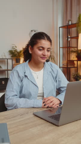 Young woman wearing headset, freelance worker, call center or support service operator helpline, talking with client or colleague communication support. Girl using laptop at home office desk. Vertical